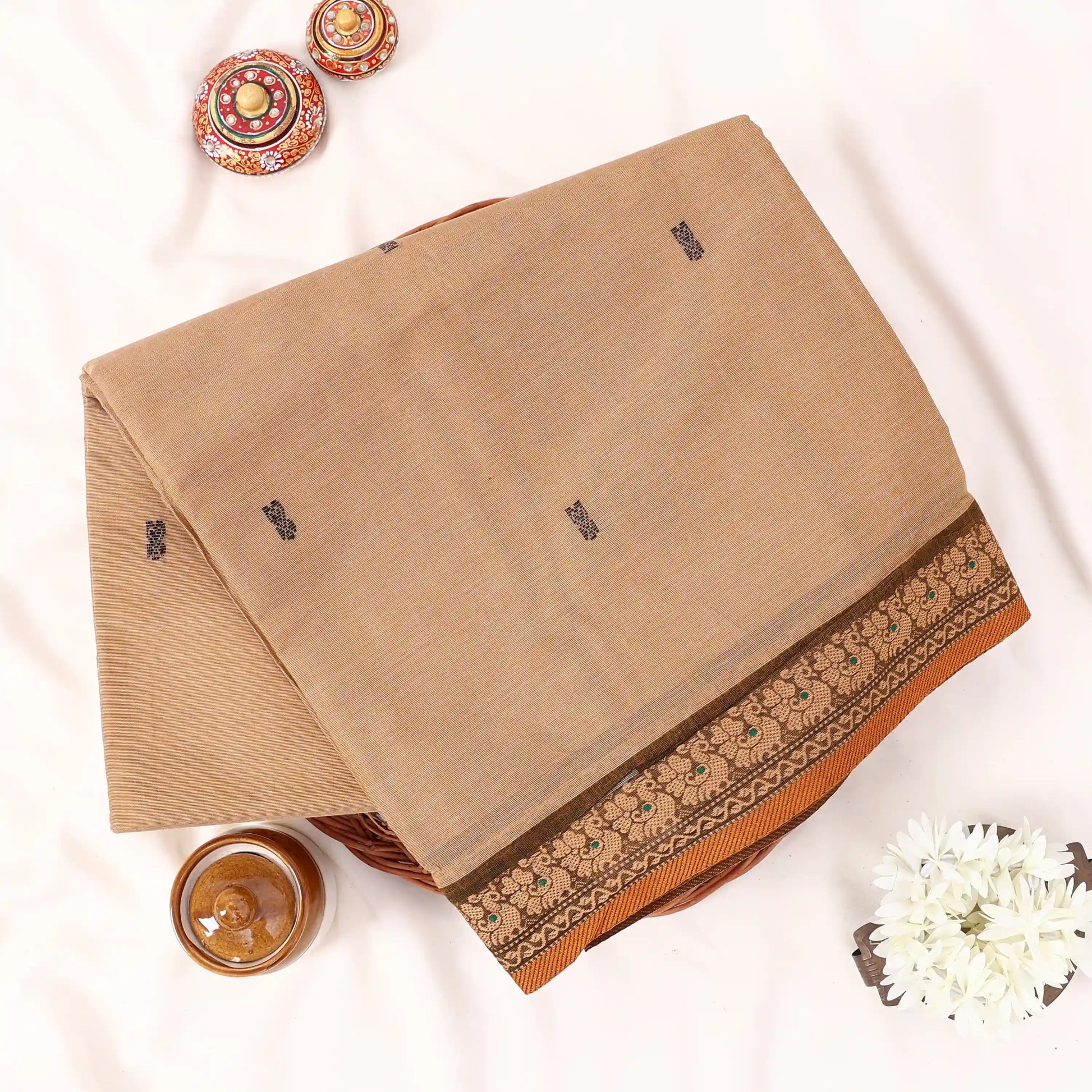 Chettinad cotton saree with beige body black buttas and brown peacock zari border folded in a basket top view.