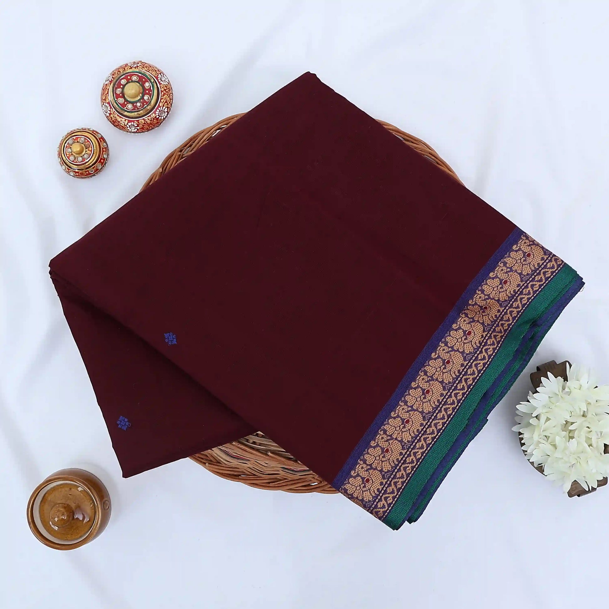 Chettinad cotton saree with maroon body purple buttas and purple peacock zari border folded in a basket top view.