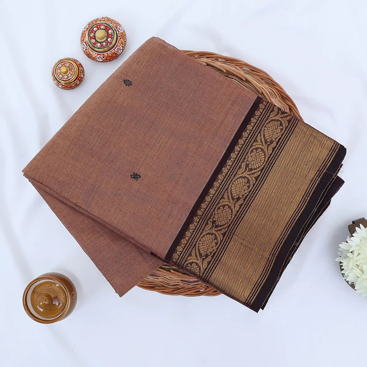 Chettinad cotton saree with brown body black buttas and gold zari border folded in a basket top view.