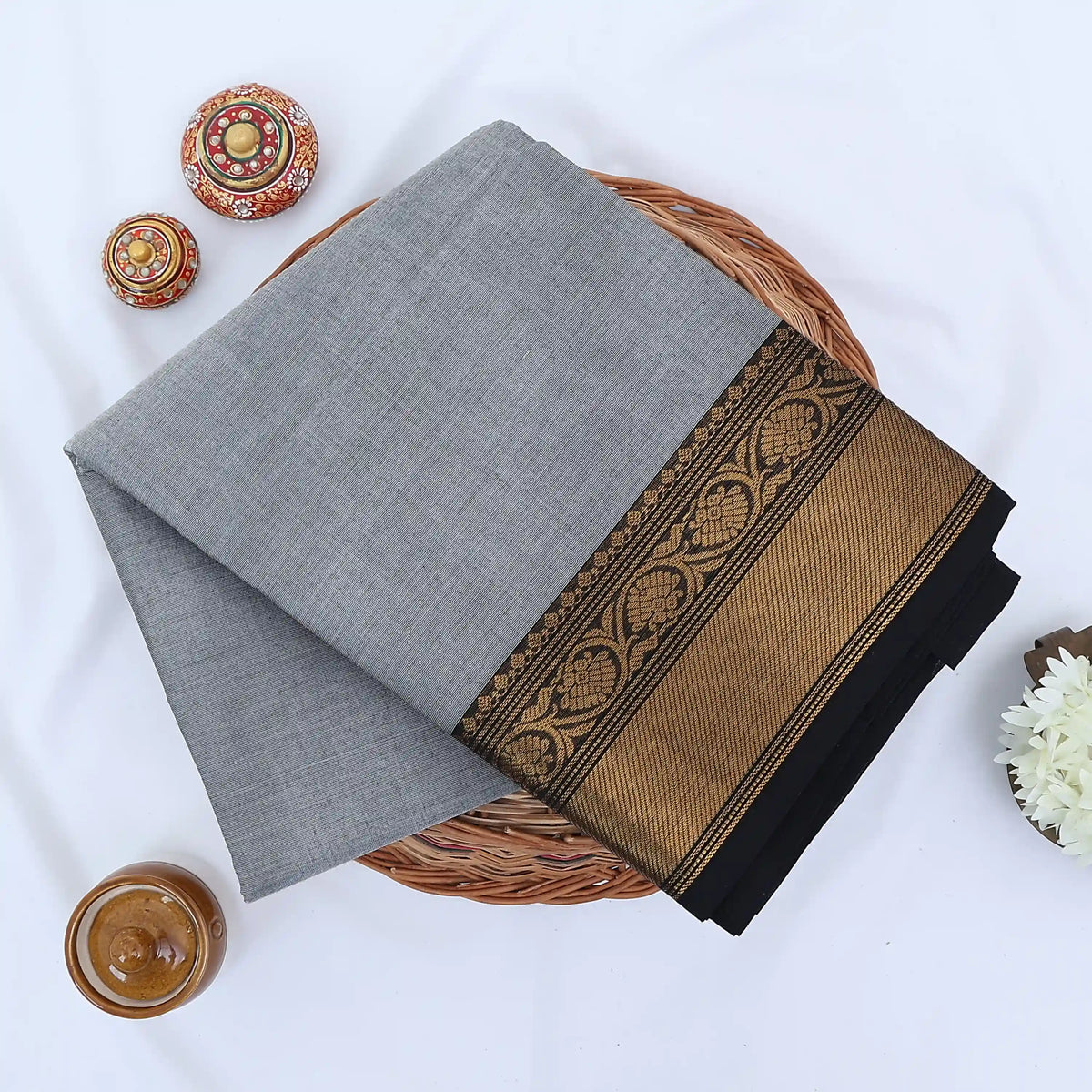 Chettinad cotton saree with grey body black buttas and gold zari border folded in a basket top view.