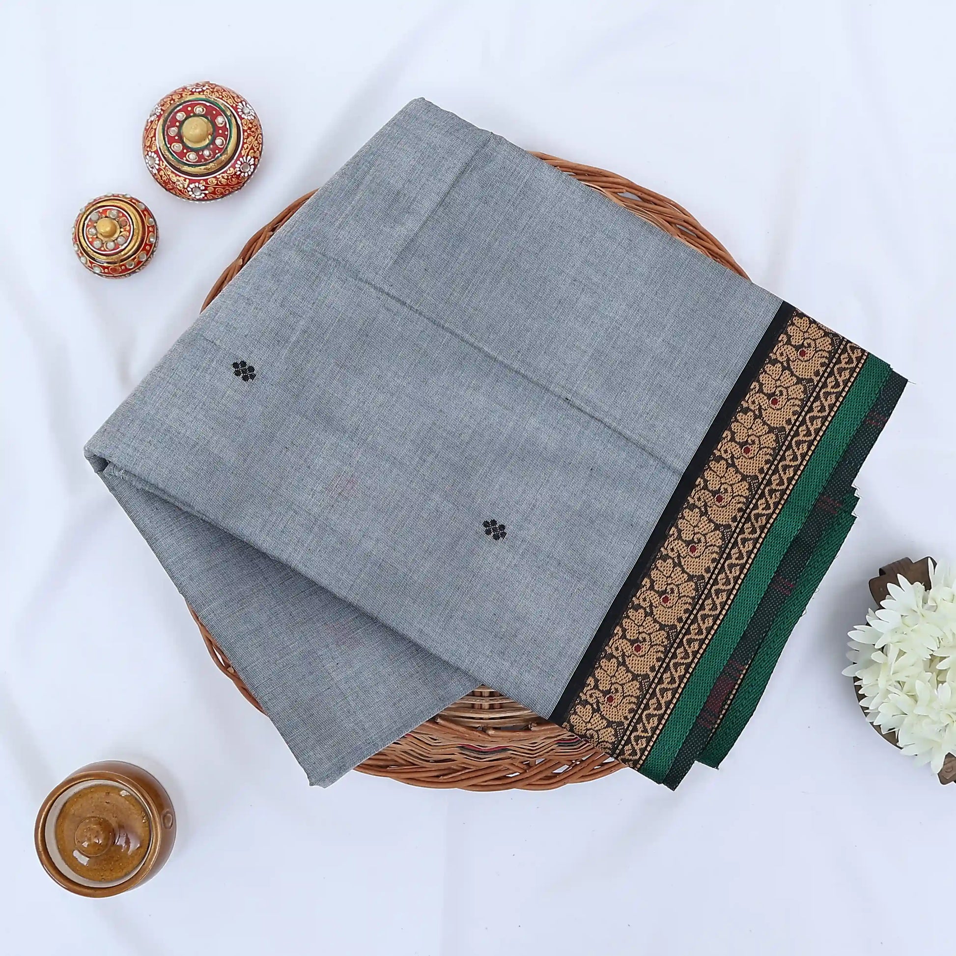 Chettinad cotton saree with grey body black buttas and black peacock zari border folded in a basket top view.