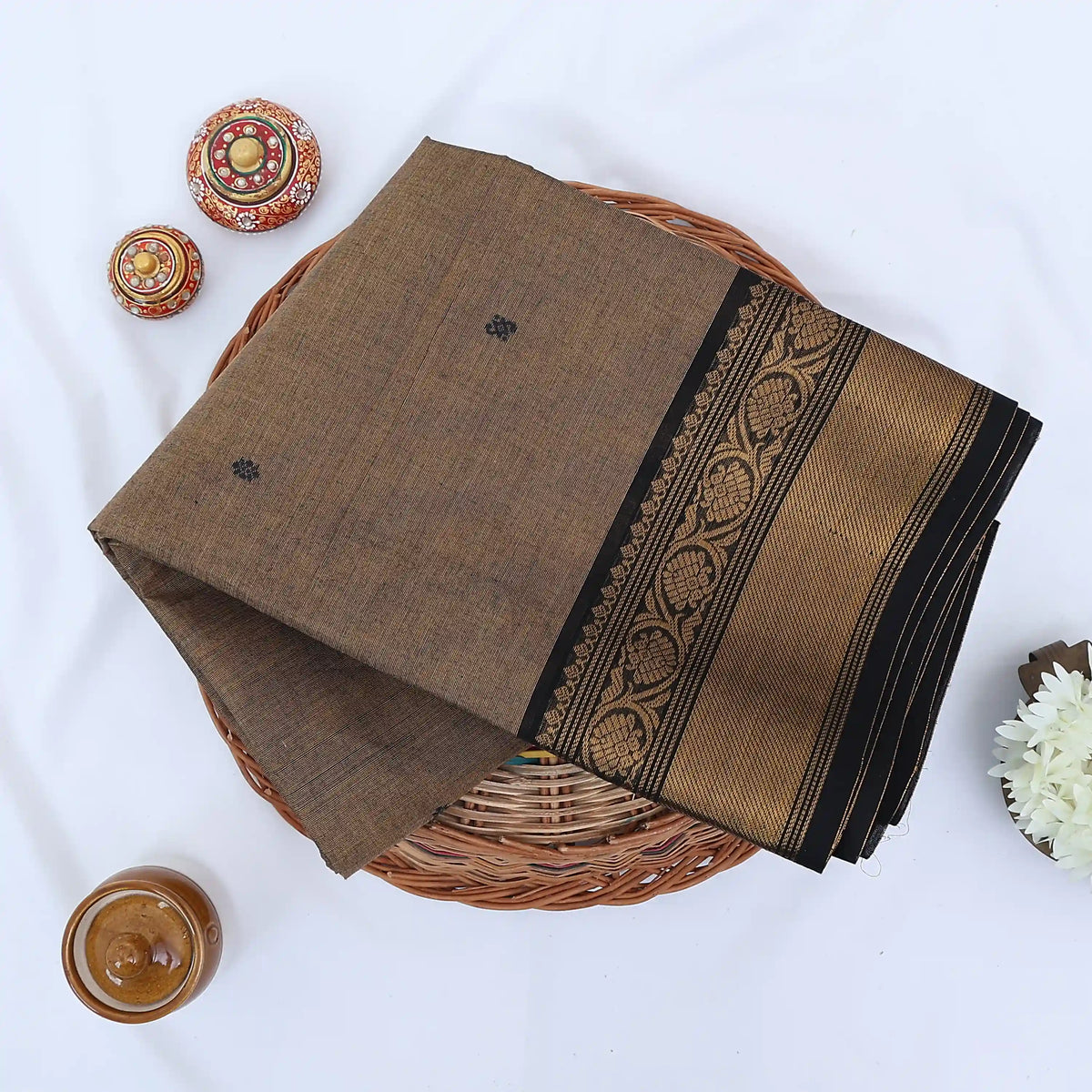 Chettinad cotton saree with light brown body black buttas and gold zari border folded in a basket top view.