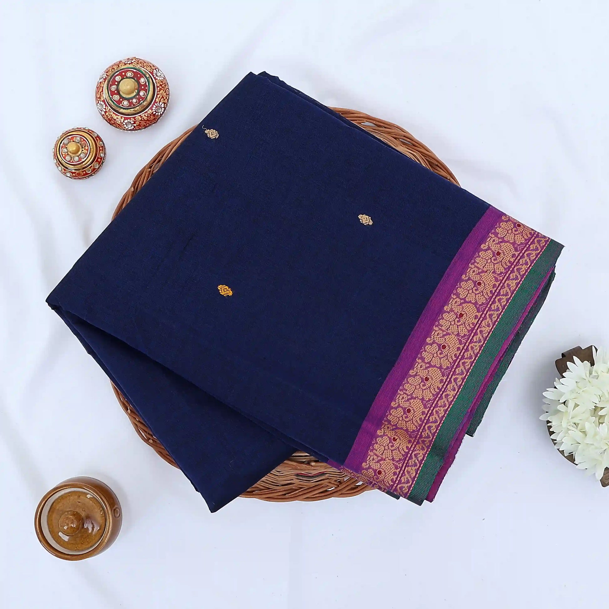 Chettinad cotton saree with navy body yellow buttas and pink peacock zari border folded in a basket top view.