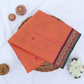 Chettinad cotton saree with orange body multi buttas and maroon peacock zari border folded in a basket top view.