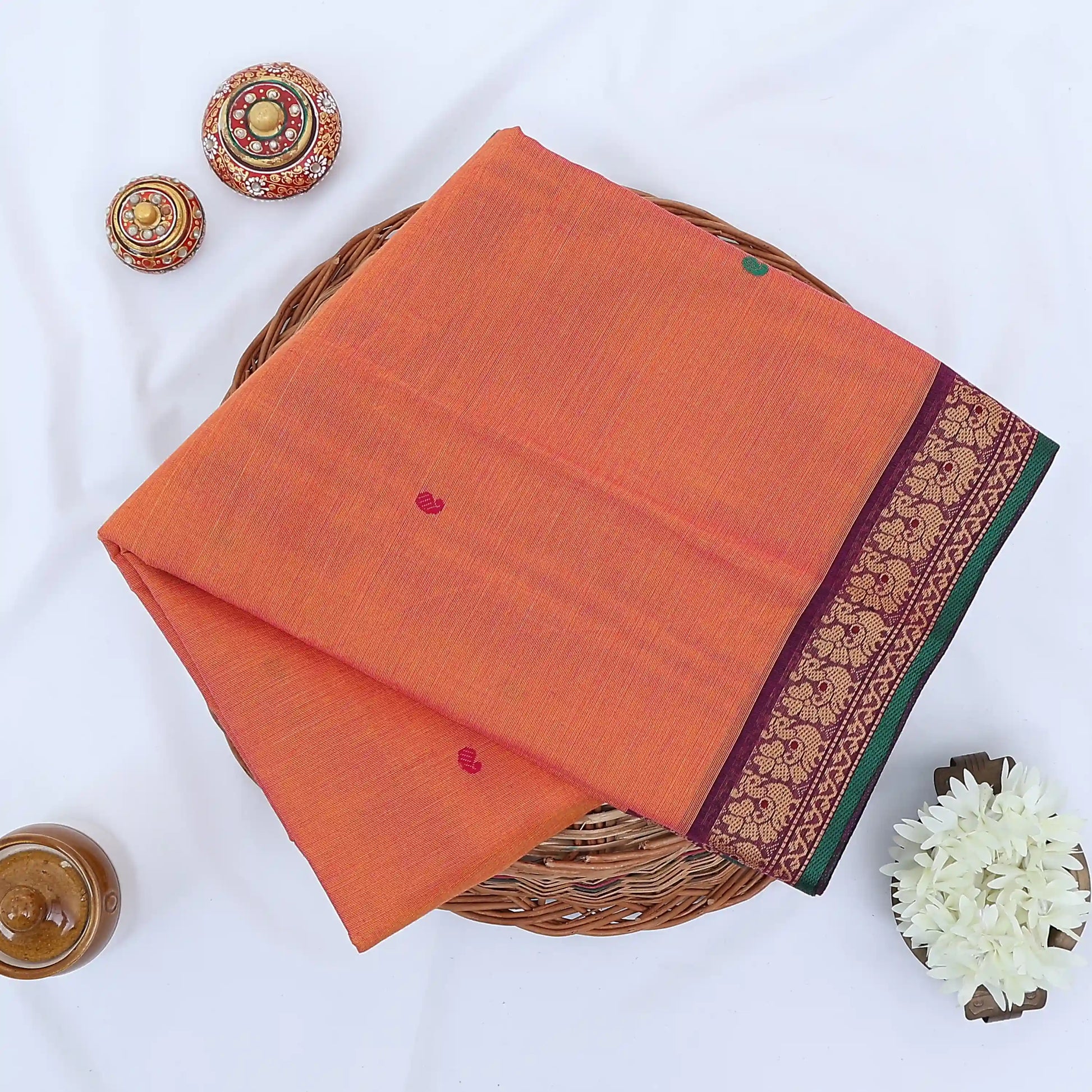 Chettinad cotton saree with orange body multi buttas and maroon peacock zari border folded in a basket top view.