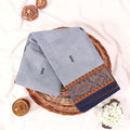 Kanchi Cotton Saree With Grey Body And Navy Gold Zari Floral Border Neatly Folded On Wicker Basket Top View
