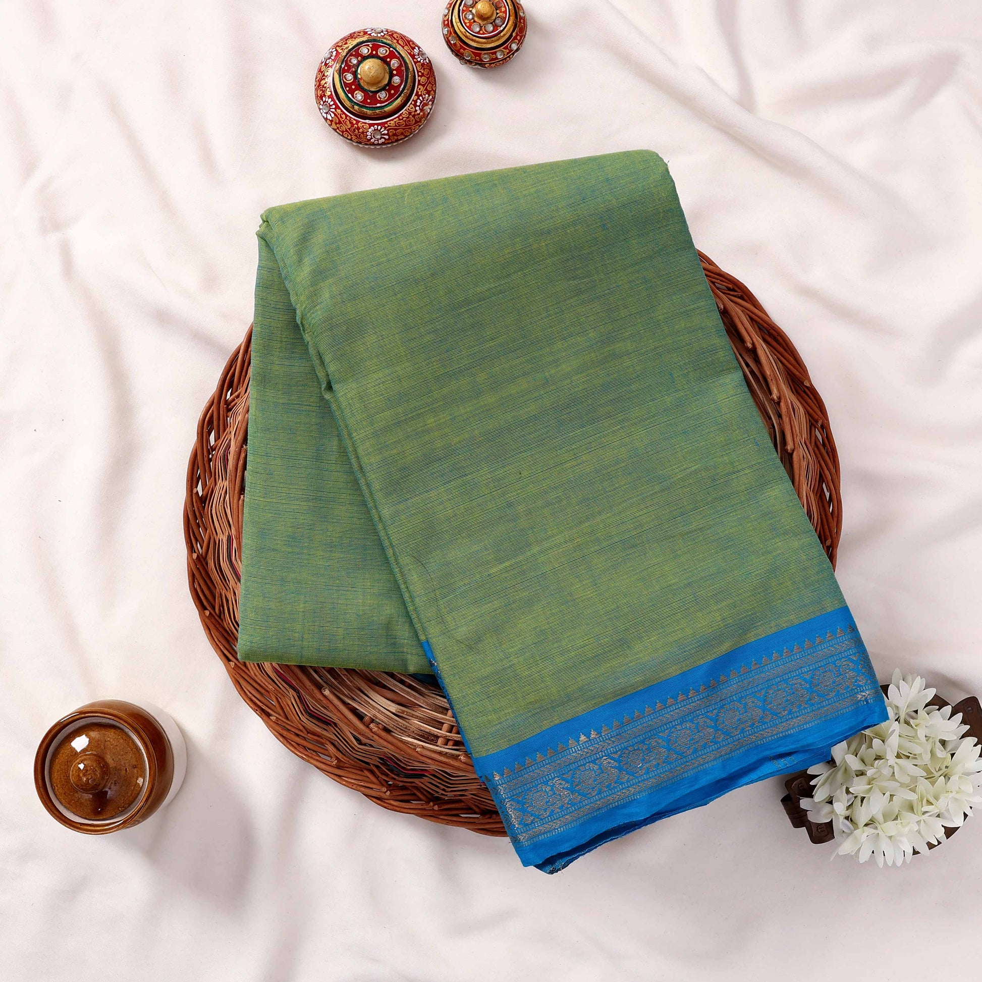 Kanchi Cotton Saree With Blue Zari Border In Light Green Folded Flat Lay View On Wicker Tray With Decor Accents
