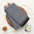 Narayanpet cotton saree with plain grey body and paisley zari woven border folded top view on wicker basket.

