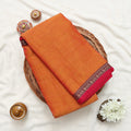 Narayanpet cotton saree with plain orange body and paisley zari woven border folded top view on wicker basket.
