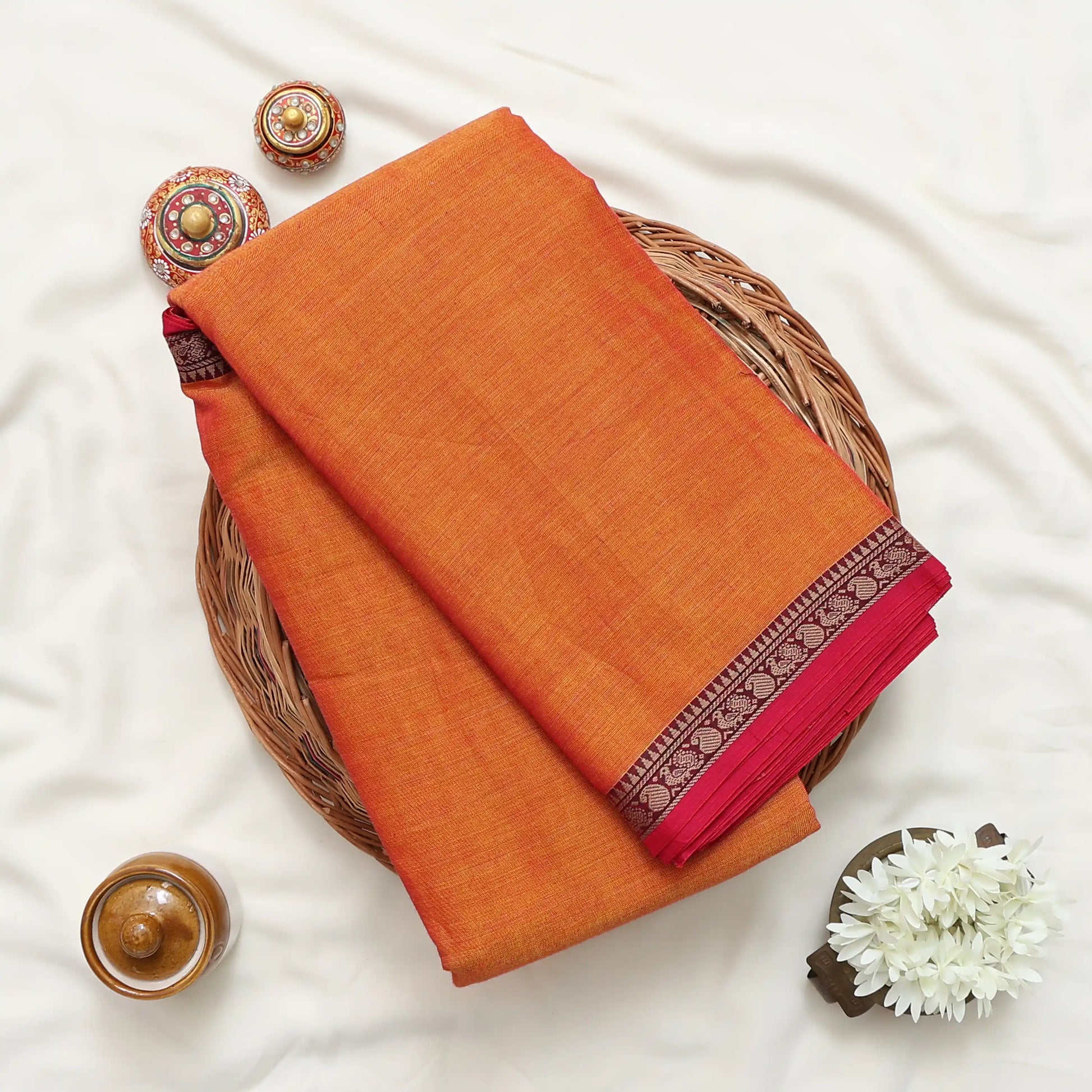 Narayanpet cotton saree with plain orange body and paisley zari woven border folded top view on wicker basket.

