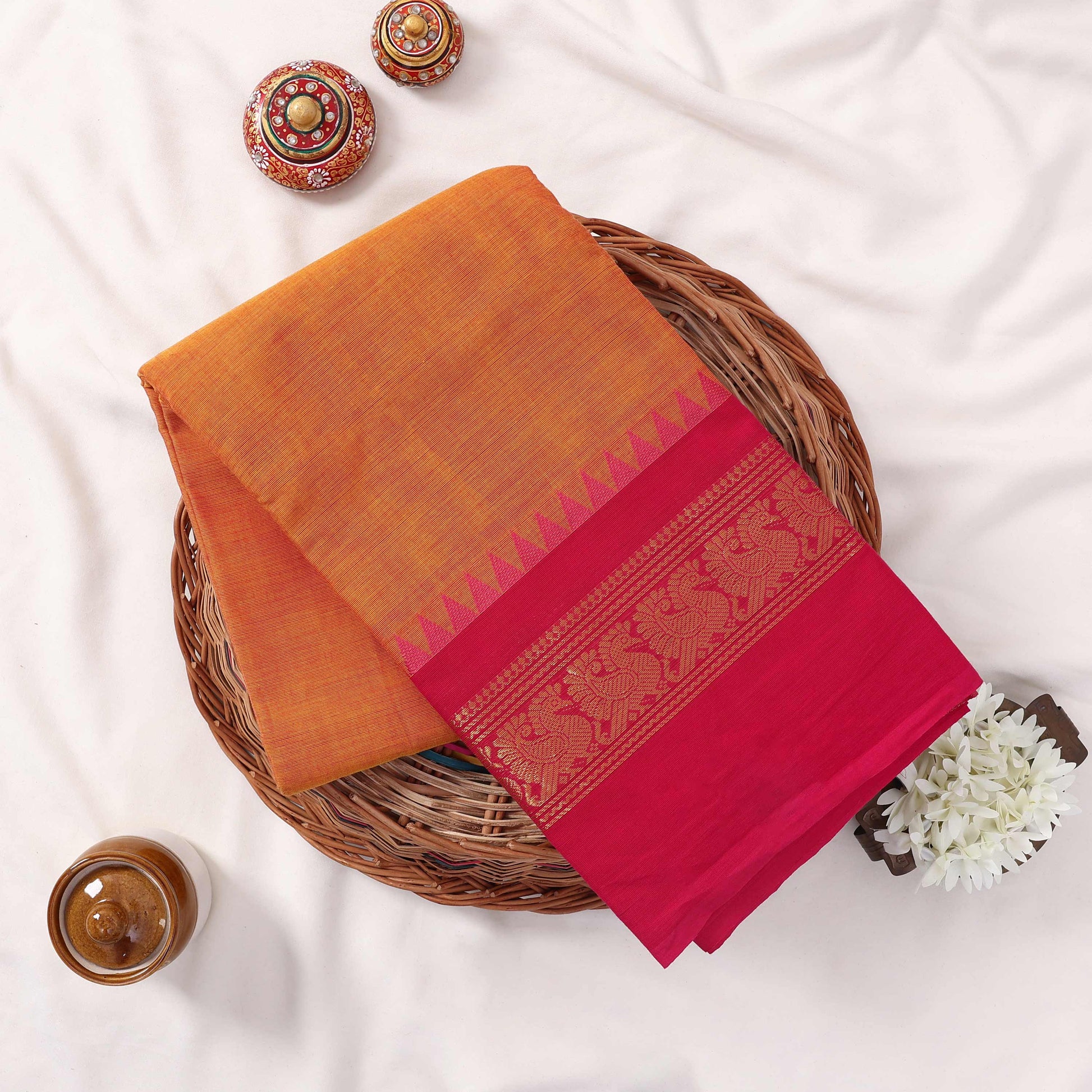 Kanchi Cotton Saree With Orange Body And Pink Zari Border Neatly Folded On A Wicker Basket With Floral Accents Top View
