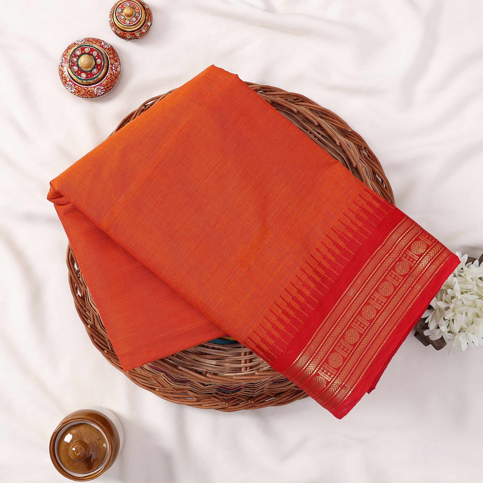 Kanchi Cotton Saree With Gold Zari Border In Vibrant Orange And Red Tones, Folded Flat Lay View With Decorative Accents
