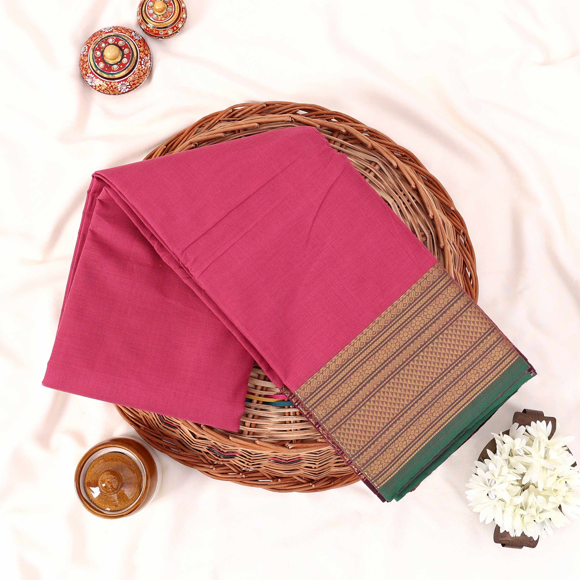 Kanchi Cotton Saree With Gold Zari Border In Pink And Green Neatly Folded On A Wicker Basket Top View Product Display
