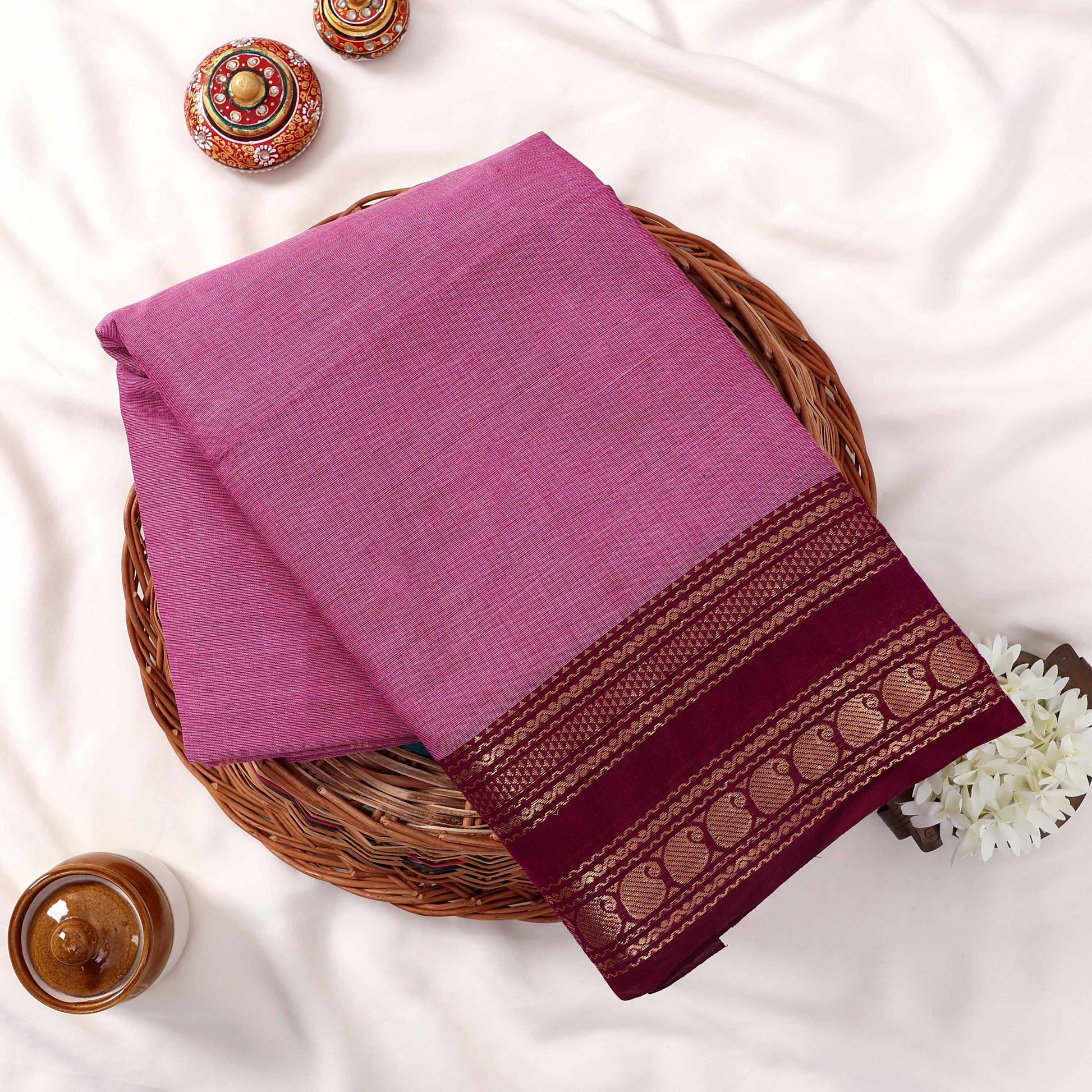 Kanchi Cotton Saree With Solid Pink Body And  Maroon Zari Border Folded Display On Wicker Basket Top View
