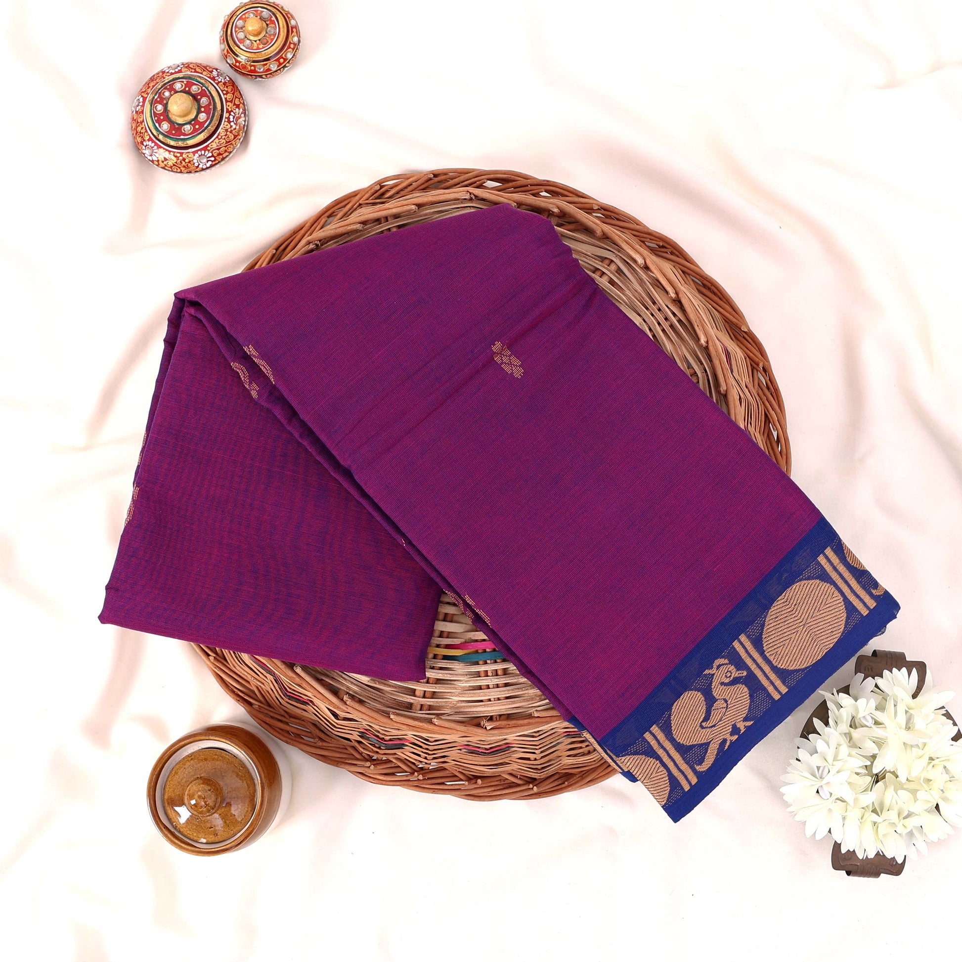 Kanchi Cotton Saree With Gold Zari Border In Vibrant Purple And Blue Neatly Folded On A Wicker Basket Top View Product Display

