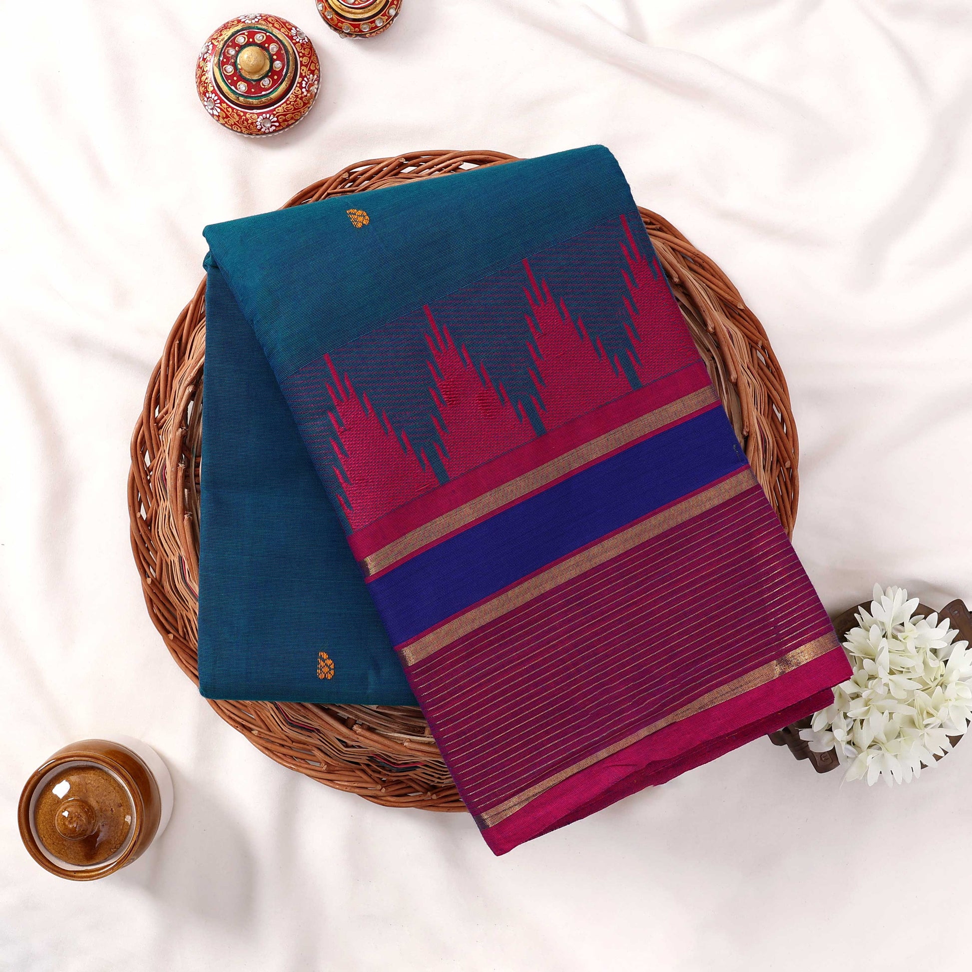 Kanchi Cotton Saree With Teal Body And Magenta Purple Zari Border Folded On Wicker Tray With Floral Décor Top View
