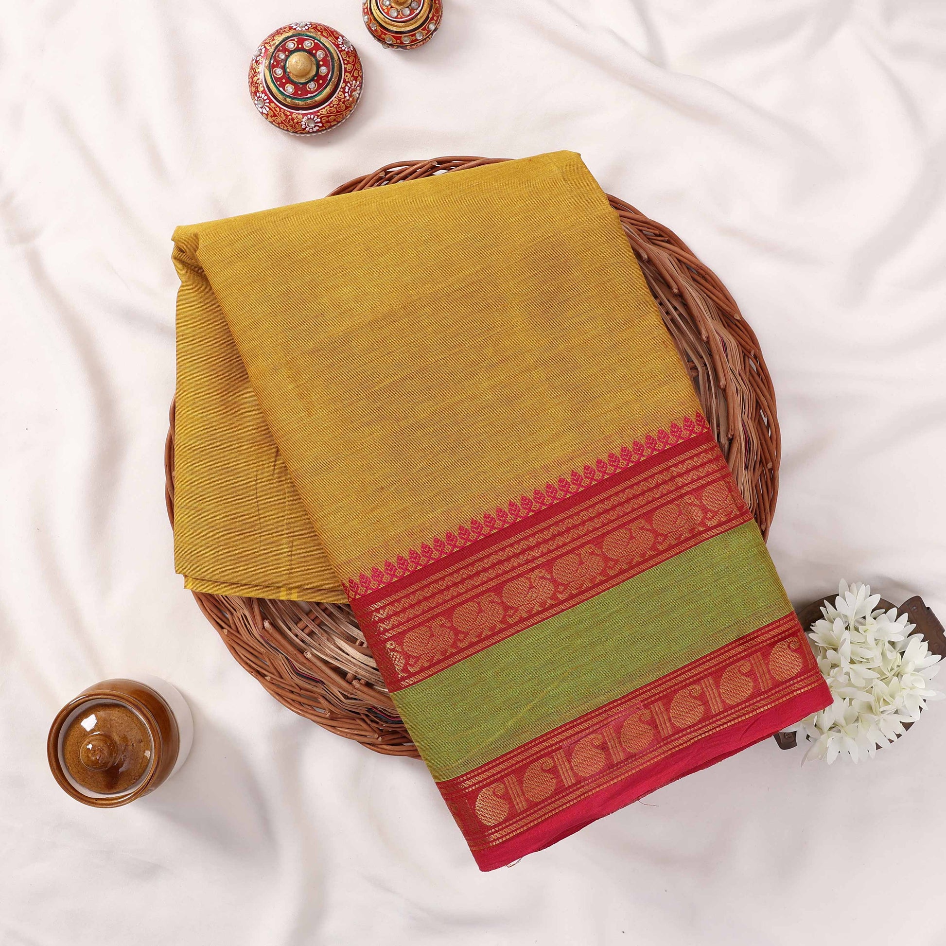 Kanchi Cotton Saree With Textured Yellow Body Green Panel And Red Zari Border Folded Flat Lay View With Decor Accents
