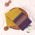 Kanchi Cotton Saree With Yellow Body And Purple Zari Border Folded On A Wicker Basket With Decor Accents Top View
