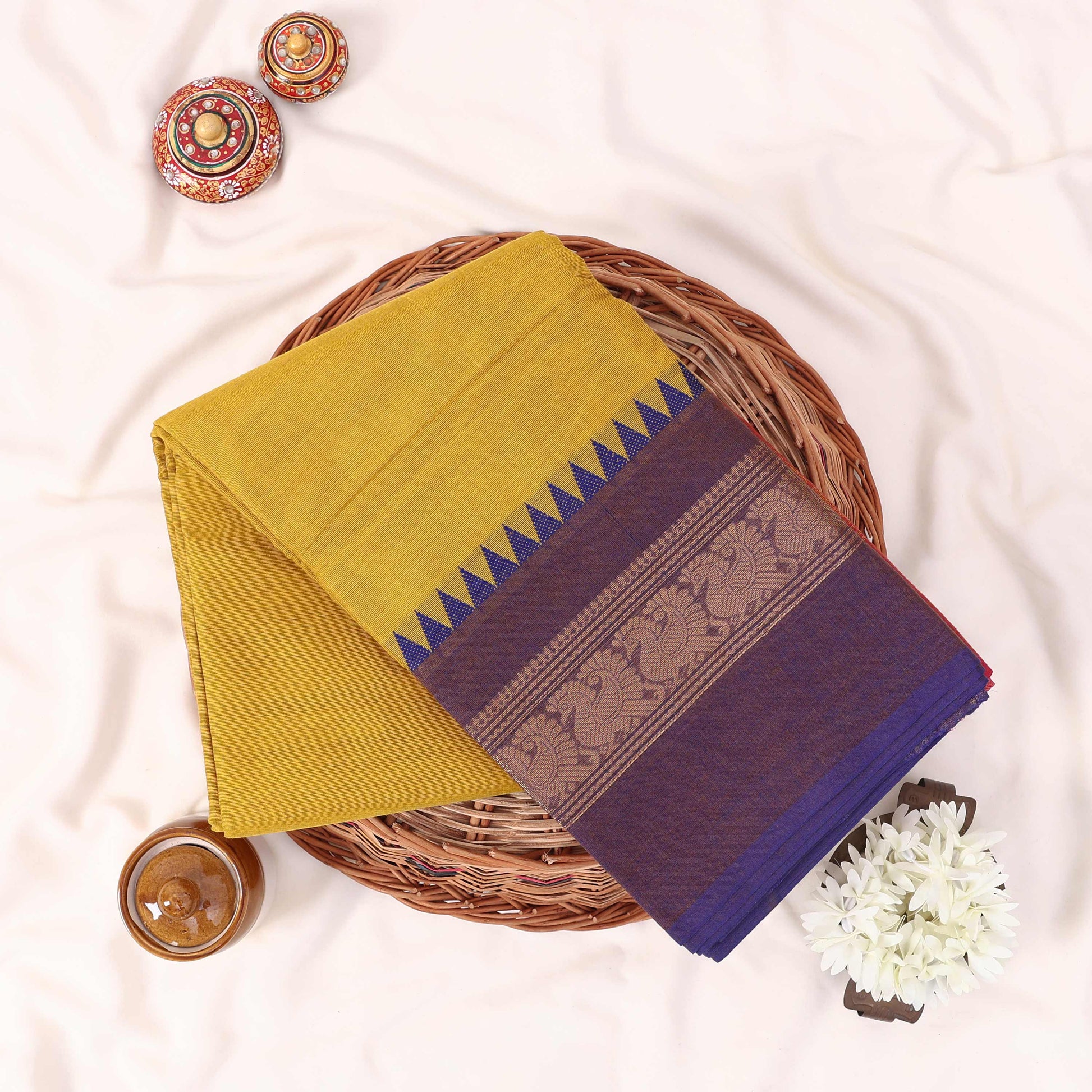Kanchi Cotton Saree With Yellow Body And Purple Zari Border Folded On A Wicker Basket With Decor Accents Top View
