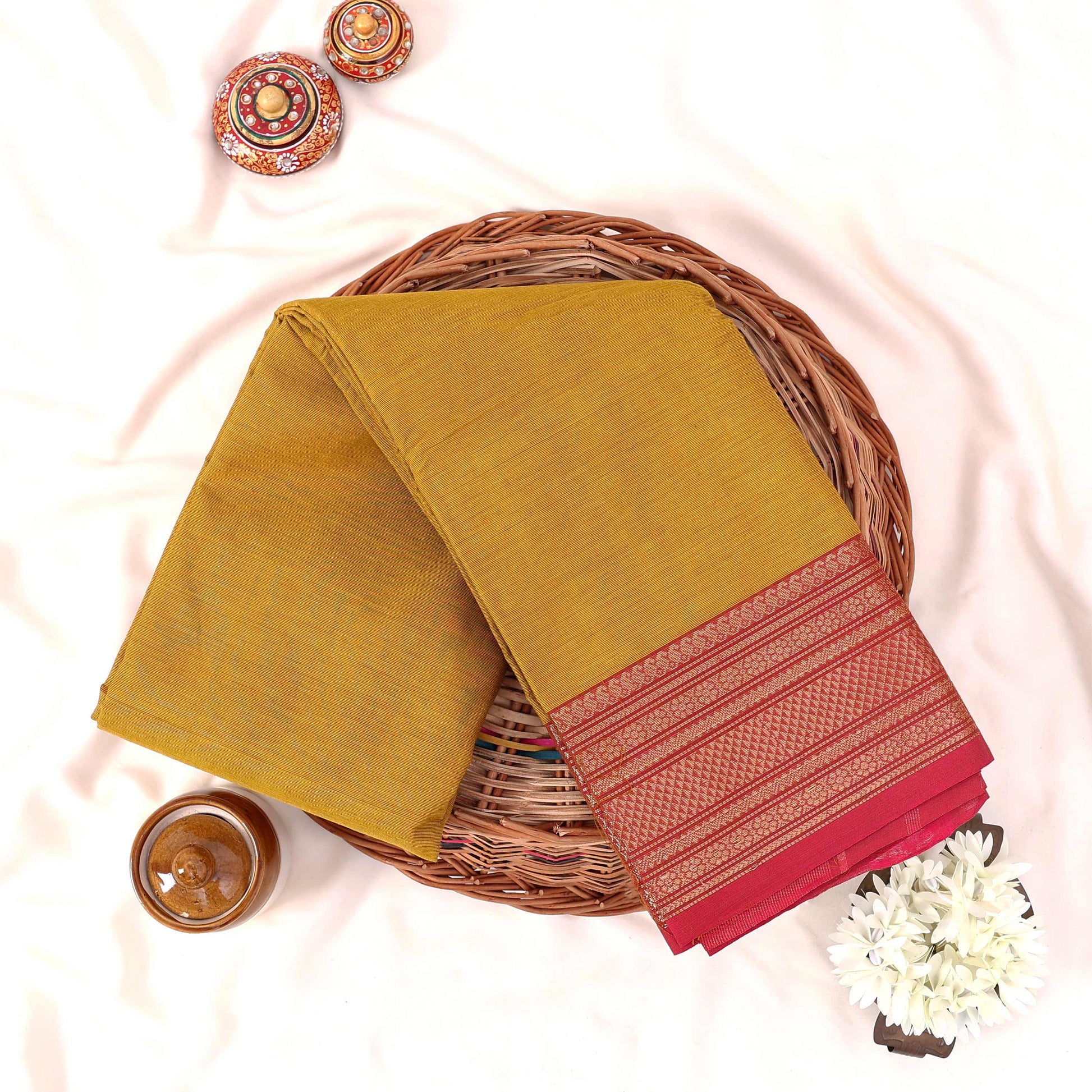 Kanchi Cotton Saree With Yellow Body And Pink Zari Woven Border Folded Flat Lay View With Decorative Basket And Floral Accents
