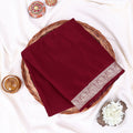 A folded deep maroon red saree,  featuring a detailed beige floral border