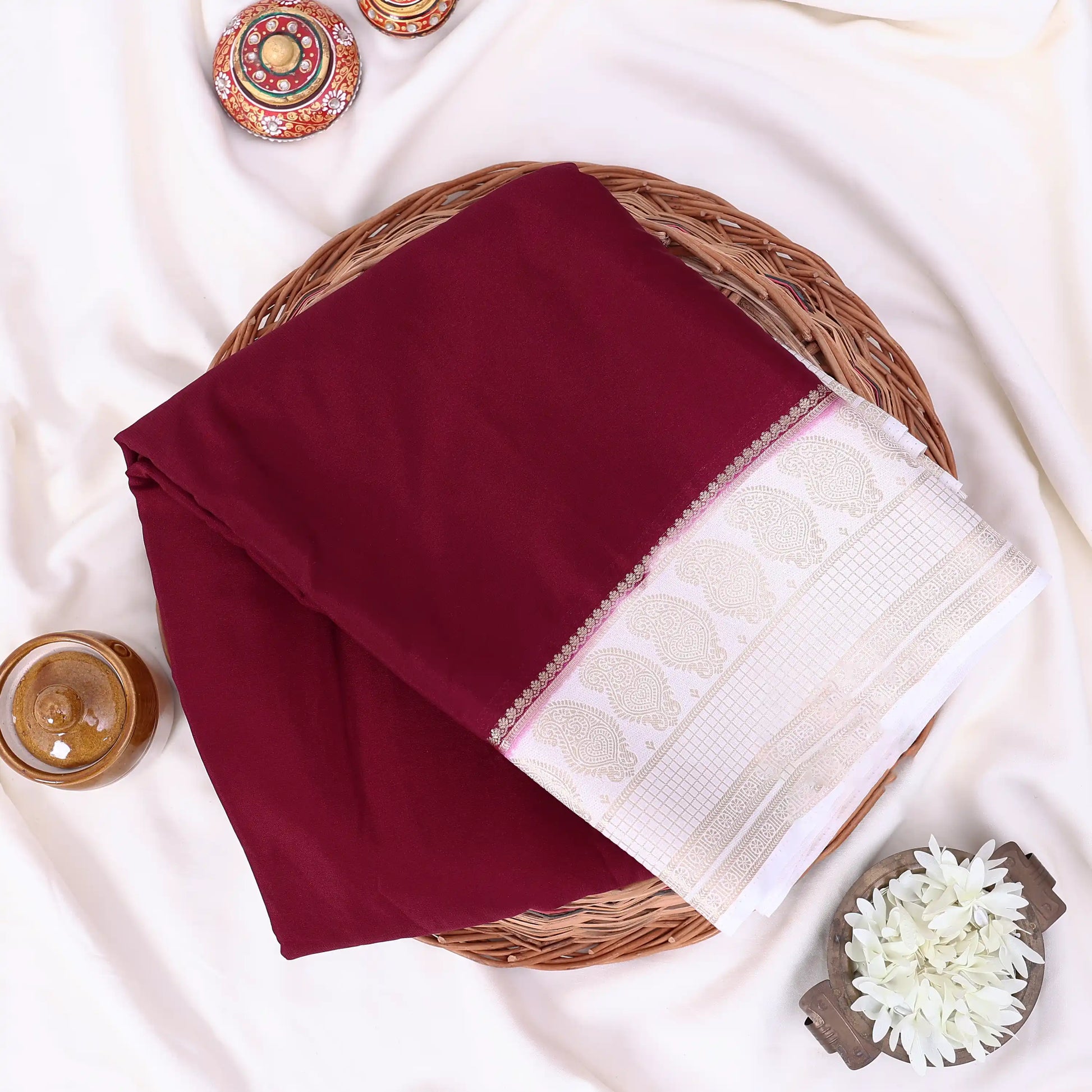 A folded deep maroon mysore silk saree features a wide white border with a beige leaf pattern and a thin pink accent line.