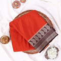 A folded orange-red mysore silk saree features a wide border with a dark brown paisley pattern and metallic silver detailing.