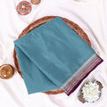 A folded light teal mysore silk saree features a wide border with a purple paisley pattern and metallic silver detailing.