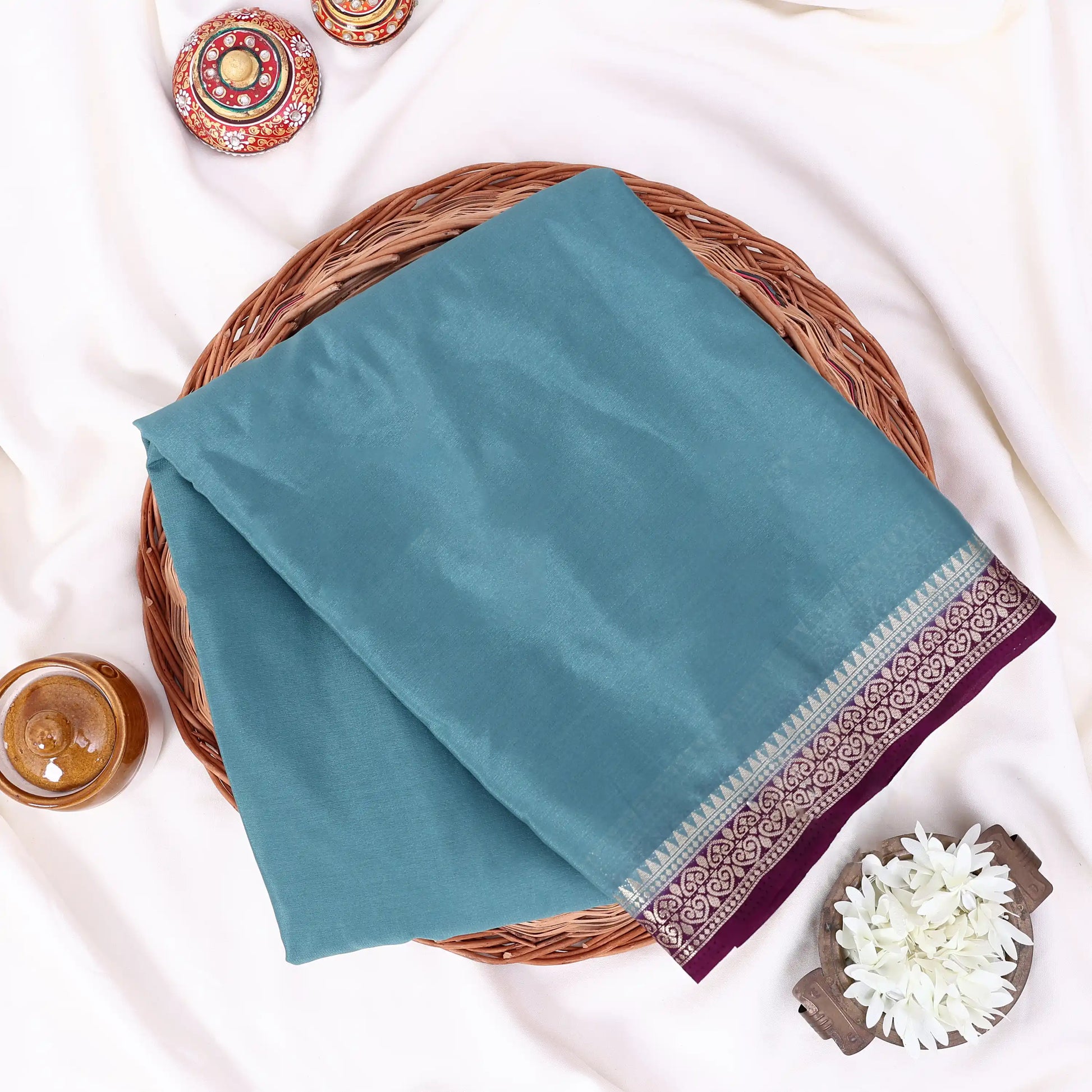 A folded light teal mysore silk saree features a wide border with a purple paisley pattern and metallic silver detailing.