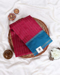 A folded silk saree has a deep magenta-pink body with fine vertical gold stripes and a contrasting teal-blue border with gold stripes.