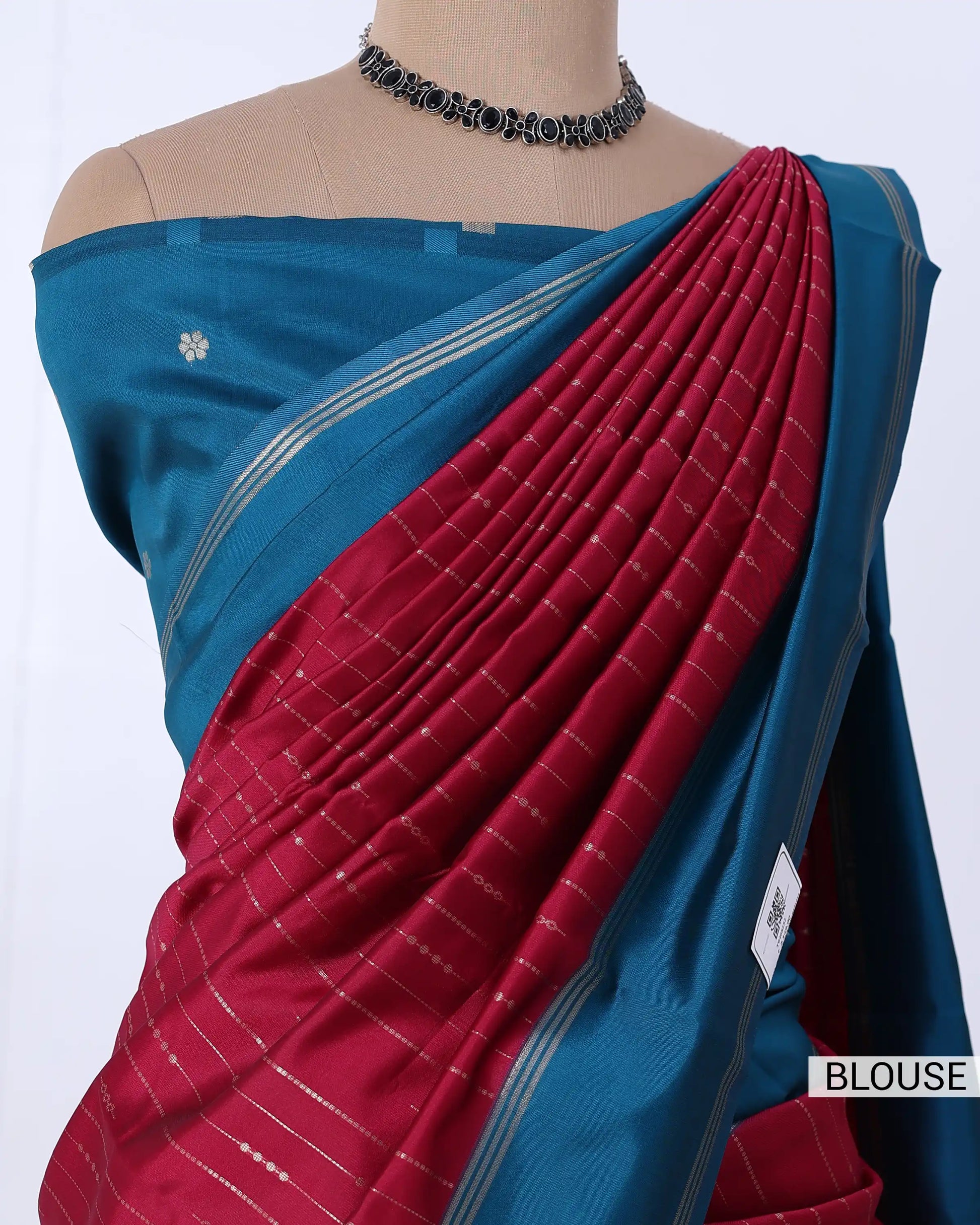 A close-up of a sari draped on a mannequin torso. The blouse portion is teal blue with a small white floral motif, and the draped pallu is deep red with fine white striped woven detailing.