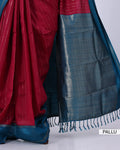 A close-up of a traditional saree fabric, showing a deep red body with fine vertical metallic zari stripes, a dark teal-blue border with woven geometric patterns, and matching blue tassels. 