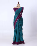 A teal and purple silk saree features a deep purple border, pallu, and blouse, with a teal body decorated with fine silver zari patterns.