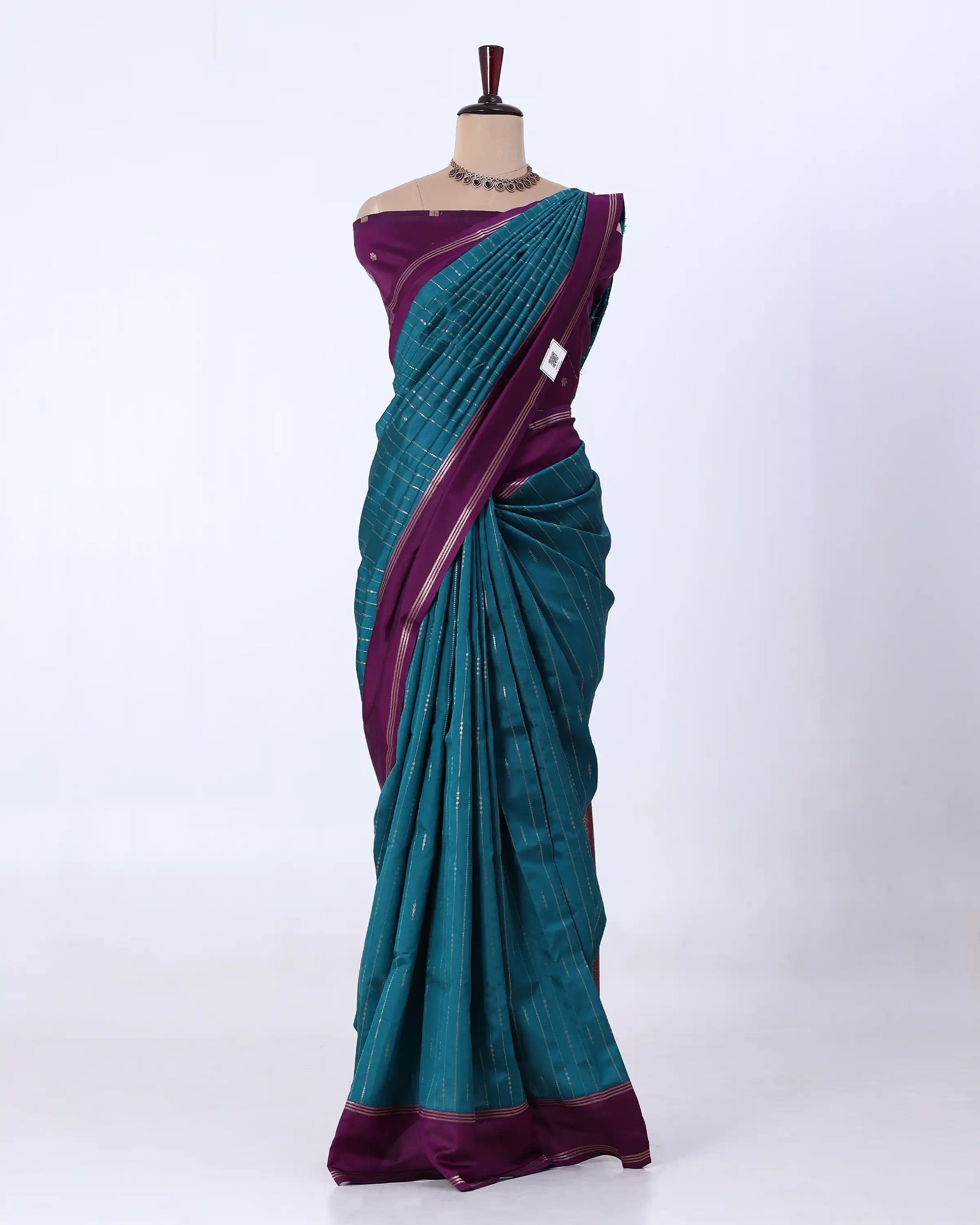 A teal and purple silk saree features a deep purple border, pallu, and blouse, with a teal body decorated with fine silver zari patterns.