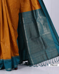 A close-up of a traditional Indian saree with a mustard yellow body, a teal blue border, and a wide teal pallu with intricate gold zari woven patterns.